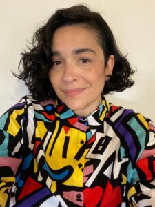 In front of a cream colored background, the poet, with chin-length dark hair wears a funky multi-colored button-down shirt.