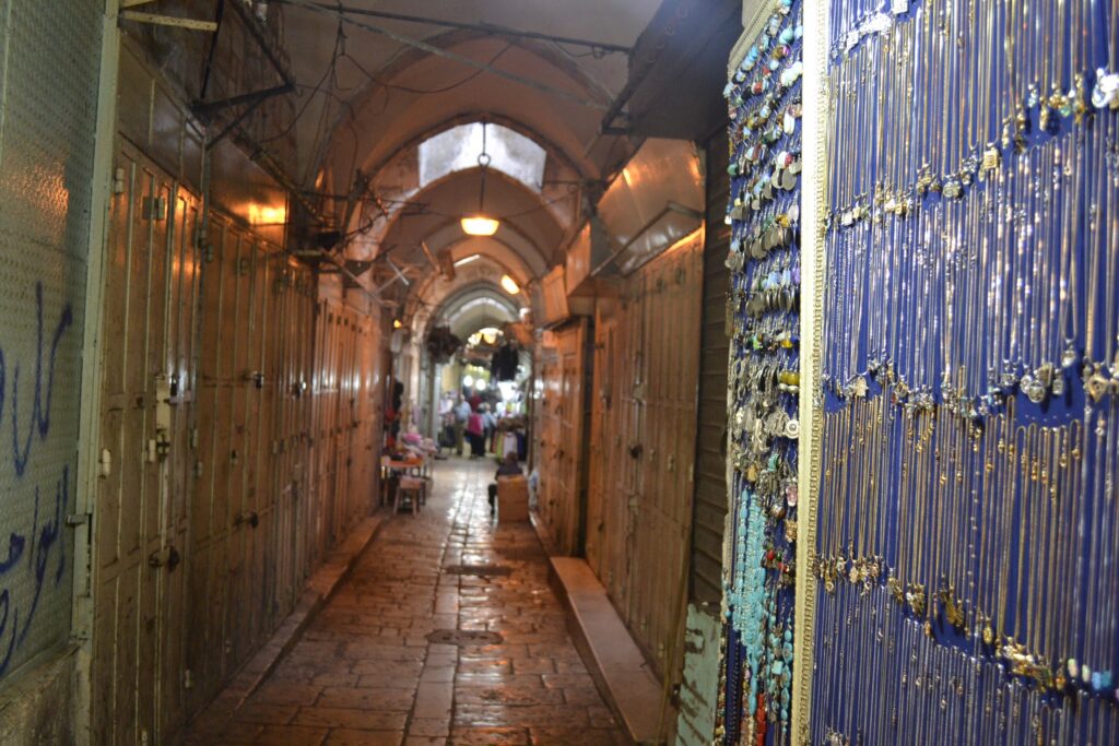 A narrow alleyway with arched ceilings and closed doors along both sides. On the right, a display of various necklaces hangs. At the far end, people are gathered at a well-lit area.