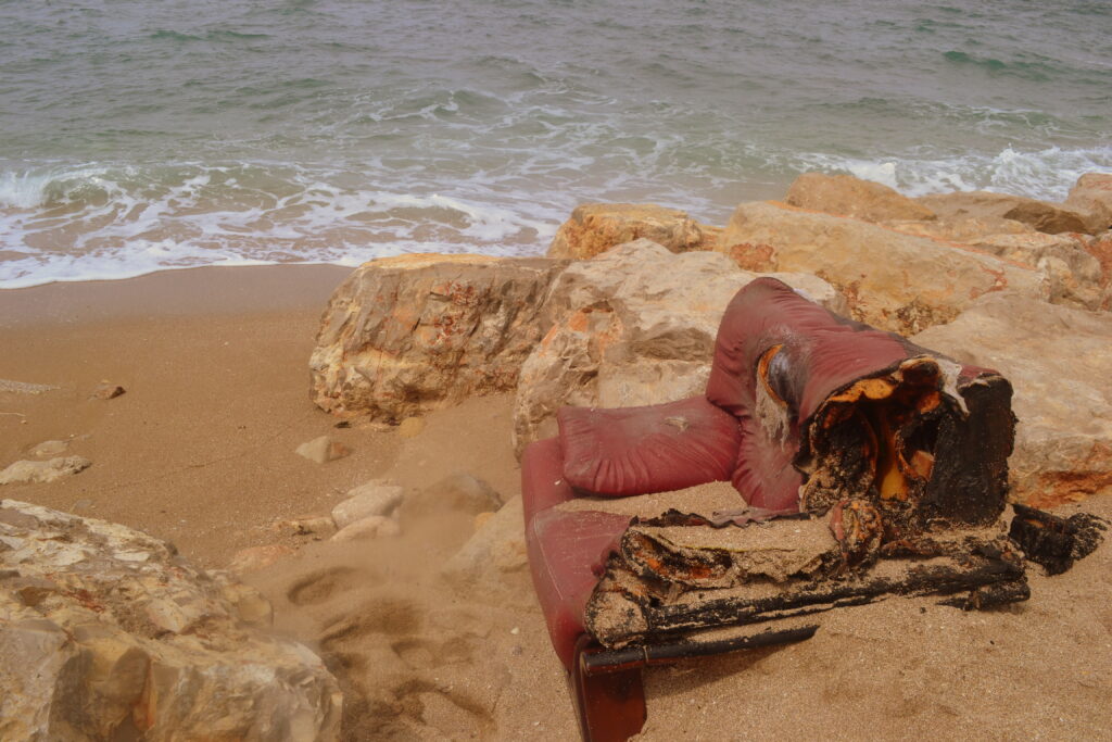 On the sandy shore of a beach, a partially burned red couch. Sea water in the background.