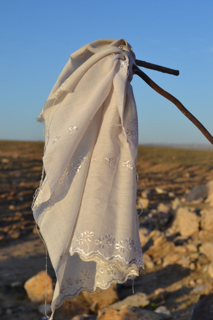 In the foreground, a torn cloth from an embroidered white veil, tied to a stick, with a rocky landscape in the background.