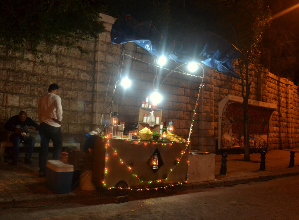 The booth of a street vendor, lit up with small multicolored lights as well as larger white lights. A man in a white shirt stands near by, hands on hips. Behind him is a tall stone wall.